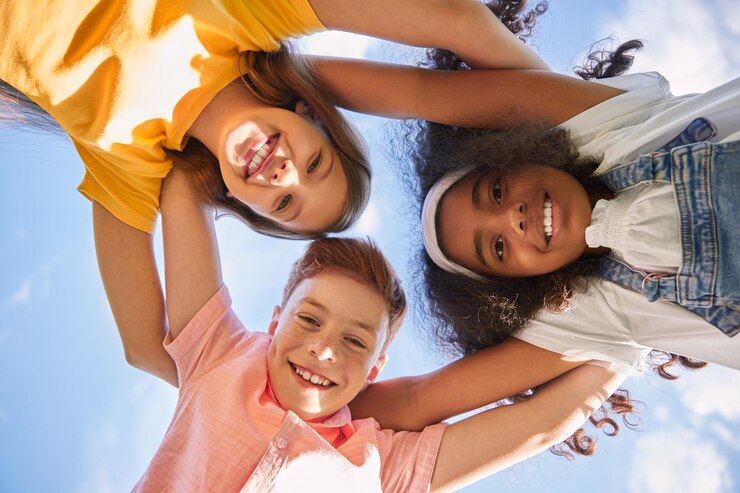 Photo from below cheerful diverse children smiling and looking at camera while standing in circle and embracing each other against cloudy blue sky