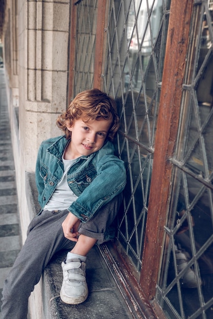 From above of cheerful kid with curly hair in denim jacket sitting on stone windowsill near tall window with metal lattice in ancient building and looking at camera