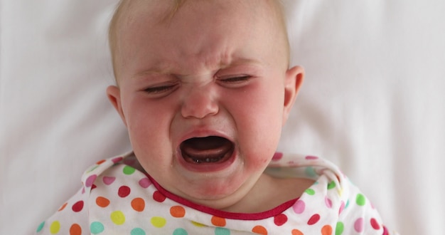 From above cute infant in colorful clothing crying while lying down on white bed sheet