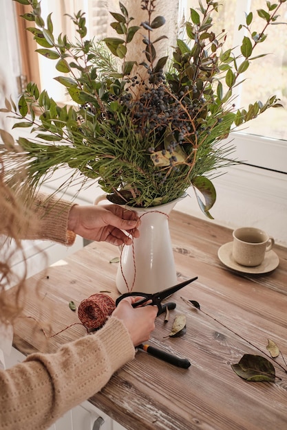 From above of faceless specialist with scissors arranging bouquet from lush green twigs in vase while working in light room