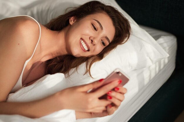 From above of happy young female lying in comfy bed with soft pillow and browsing mobile phone after awakening in morning