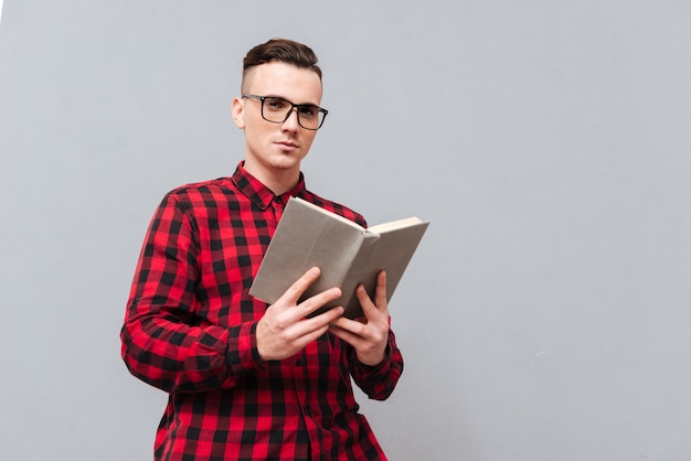 From below image of young serious man in glasses and red shirt reading book in studio. Isolated gray background