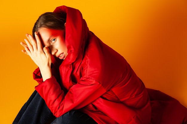 From above lonely female model with creative makeup wrapping in red oversize coat and looking away against yellow background