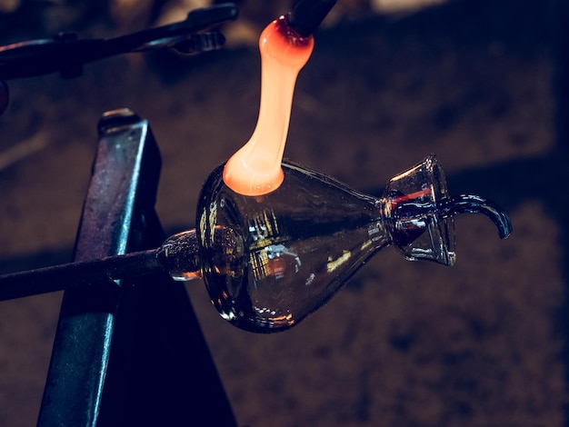 From above of master making glass handle for glass carafe fixed on metal bench using hot ductile glass and tools in glass factory