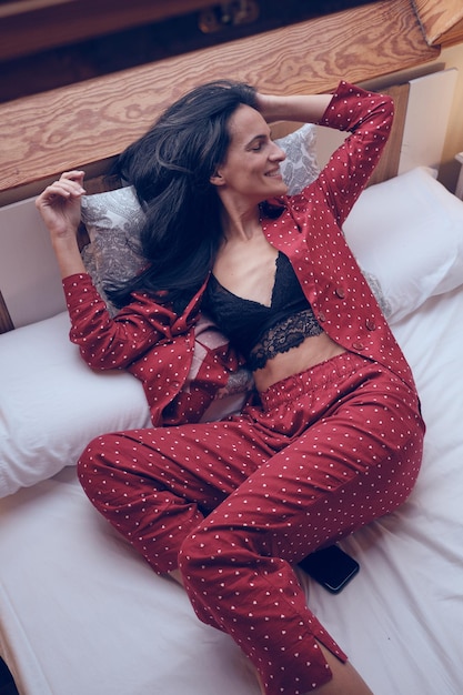 Сверху позитивная женщина в стильной пижаме трогает волосы, лежа на белой кровати в светлой спальне дома