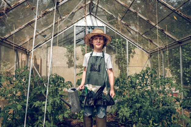 From below of positive preteen boy gardener in wicker hat and apron standing in hothouse among green plants with watering can