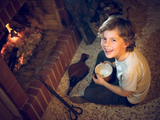 From above of preteen boy sitting with crossed legs and empty mug opposite burning fireplace