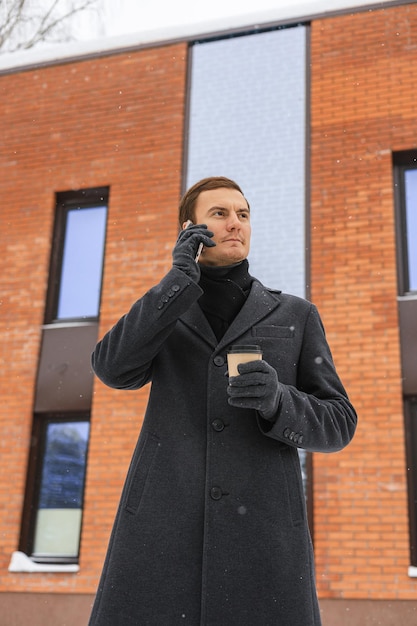 From below of serious businessman in coat with cup of takeout coffee making phone call on smartphone