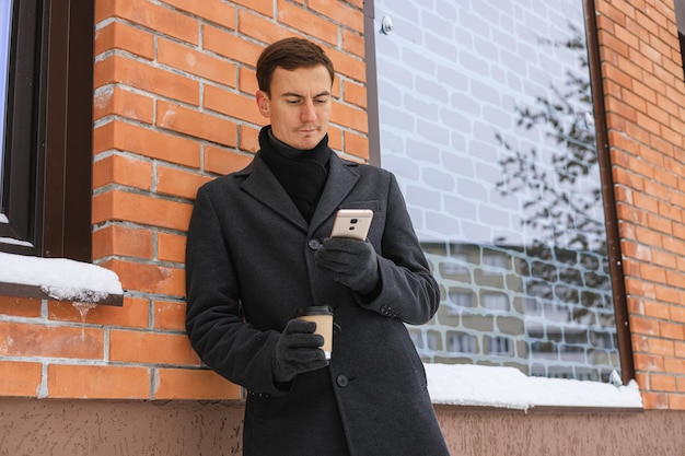 Снизу серьезный мужчина-предприниматель в зимнем пальто с чашкой кофе просматривает смартфон