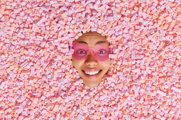 From above shot of cheerful ethnic female model smiles broadly covered with delicious sweet pink marshmallow wears trendy heart shaped sunglasses