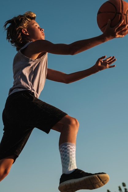 From below side view of energetic boy throwing ball while playing basketball against blue sky