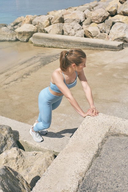 From above side view of female athlete in sportswear leaning with hands on stone fence while warming up and looking away