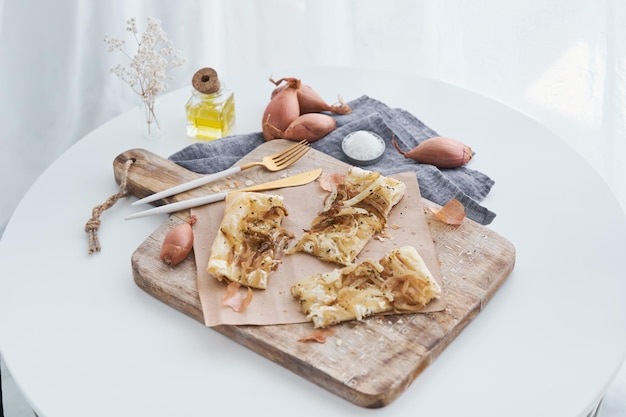 Сверху кусочки вкусного пирога из слоеного теста с луком подаются на деревянной разделочной доске со столовыми приборами на кухне