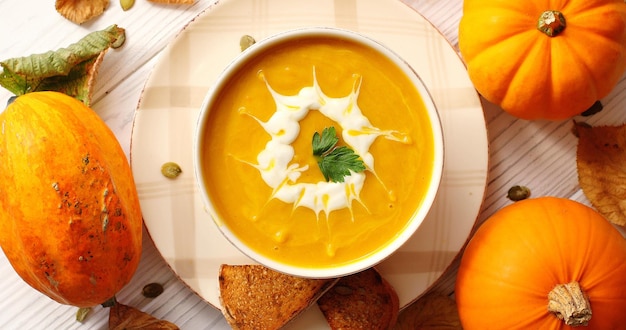 From above view of creamy yellow pumpkin soup in bowl with bread and pumpkins placed around on wooden background