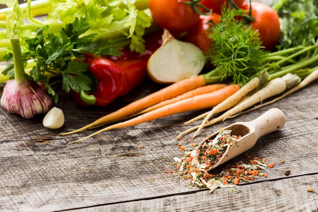 From above view wooden scoop with assortment of dry and milled vegetables spread inside and outside Large pile of fresh vegetables ingredients for condiment placed behind