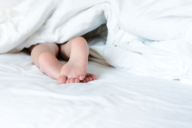 From under a white blanket, stick bare children's feet. Copy space. Bright photo in bed.