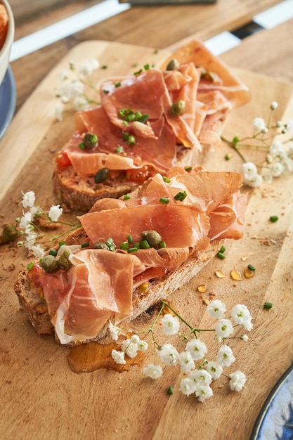 From above of yummy toasts with appetizing prosciutto and capers and herbs served on wooden cutting board and decorated with delicate white flowers on restaurant terrace