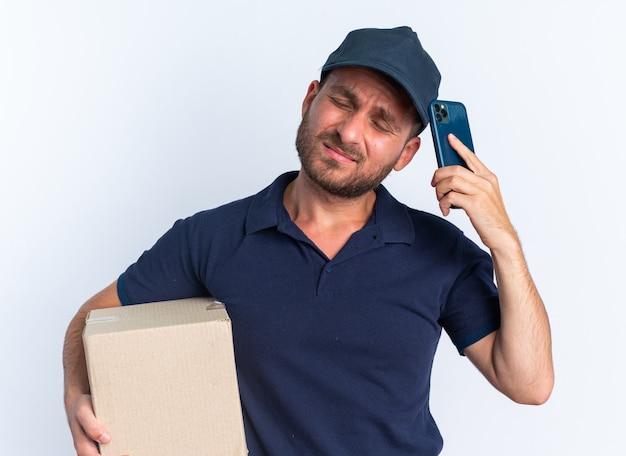 Fronsende jonge blanke bezorger in blauw uniform en pet met kartonnen doos die het hoofd aanraakt met mobiele telefoon met gesloten ogen geïsoleerd op een witte muur