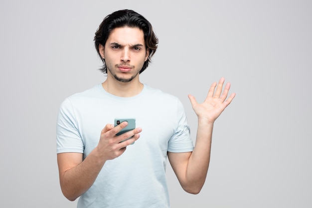 Fronsende jonge knappe man met mobiele telefoon kijkend naar camera met lege hand geïsoleerd op een witte achtergrond