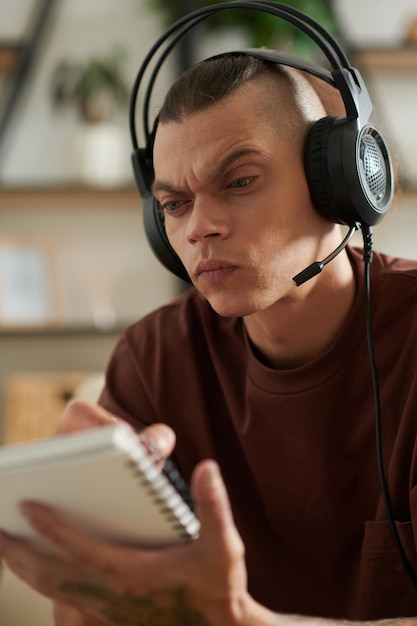 Fronsende jonge man die een headset draagt bij het kijken naar video op de computer en het opschrijven van details