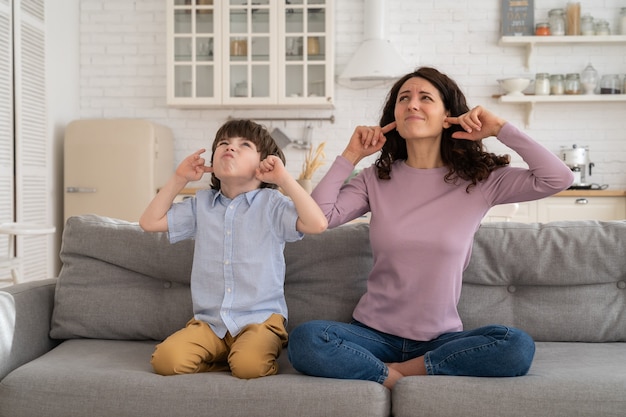 Fronsende moeder en zoon zitten op de bank en bedekken hun oren van luidruchtige muziek