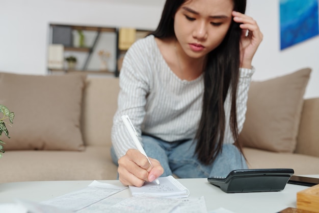 Fronsende mooie jonge Aziatische vrouw die de som van de maandelijkse uitgaven en energierekeningen telt en aantekeningen maakt in een notitieboekje