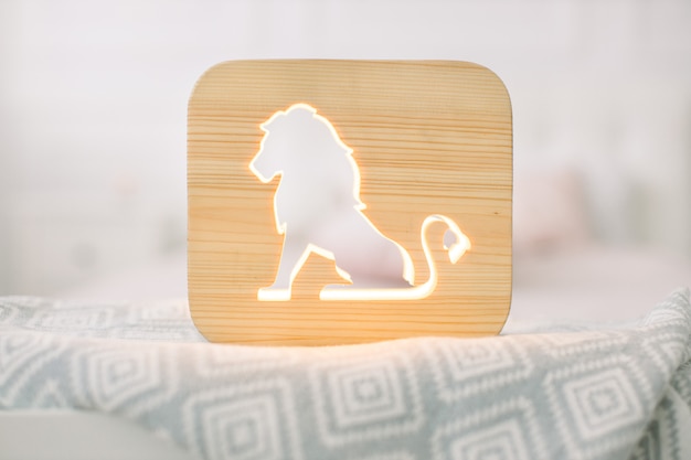 Front close up view of stylish wooden night lamp with lion cut out picture, on gray blanket at cozy light bedroom interior.