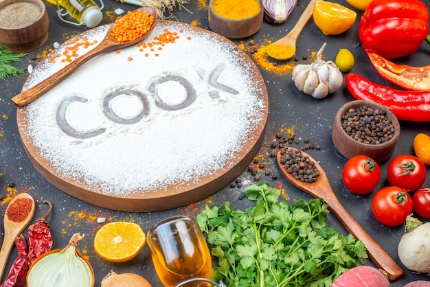 Front close view of cook writing with flour on wooden board among fresh vegetables spices green bundles fallen oil bottle on black surface