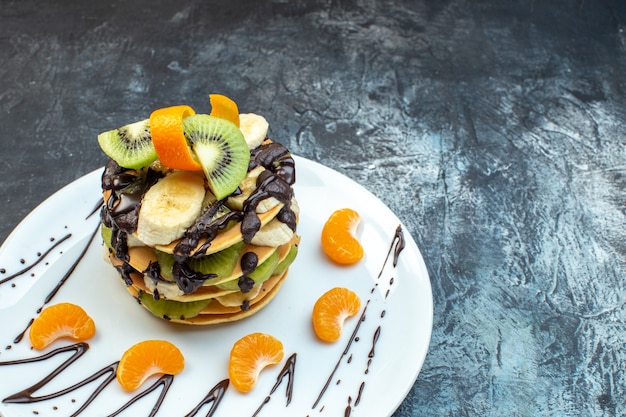 Front close view of fluffy American-style pancakes made with natural yogurt and stacked with layers of fruit decorated with chocolate on white plate on ice background