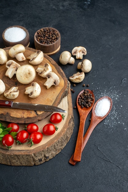 Front close view of fresh raw mushrooms and green bundle knife tomatoes spices on wooden board towel on black background