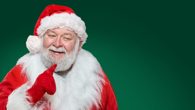 Front image of a happy Santa Claus dressed in a red coat, gloves and a hat, pointing his index finger isolated green background.