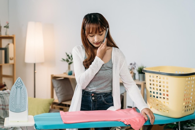 front portrait pretty asian young wife is talking on the phone while standing by the board ironing clothes in a cozy living room at home.