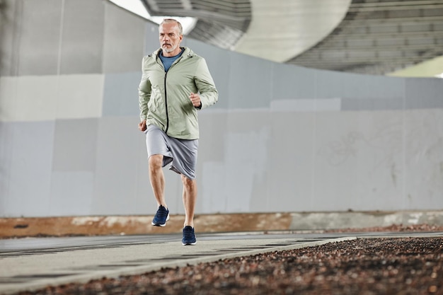 Photo front view of active mature man running towards camera outdoors against concrete background in urban