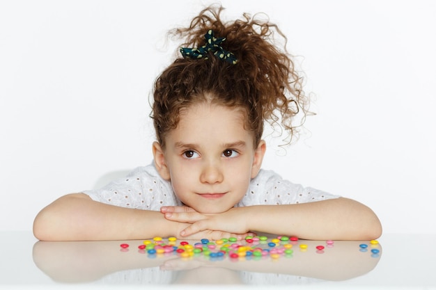 Front view of an angry little girl looking at candy hesitate to eat sitting at the table on a white background