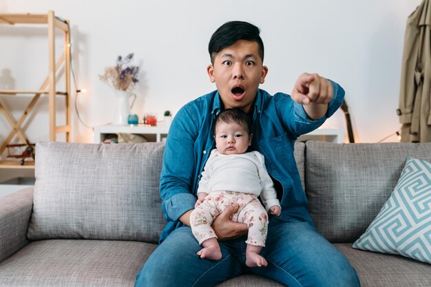 Front view asian father is finger pointing ahead with a shocked\
expression while watching tv with his innocent infant girl sitting\
in his arms on sofa at home.