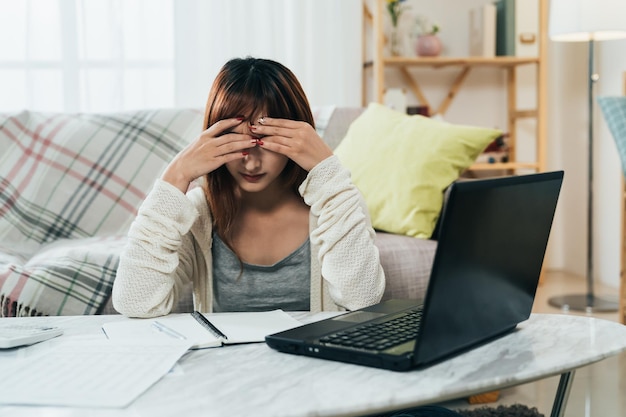 正面図アジアの女の子が座って手に頭を埋めているのは、自宅の居間でノートパソコンを使って税金を申告している間、借金を心配している.