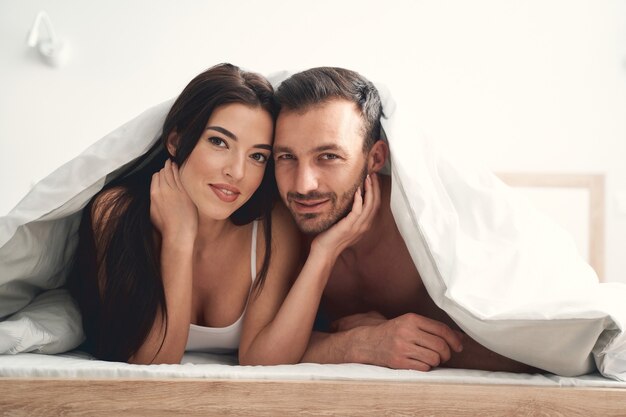 Front view of an attractive man and his wife resting in bed under the blanket