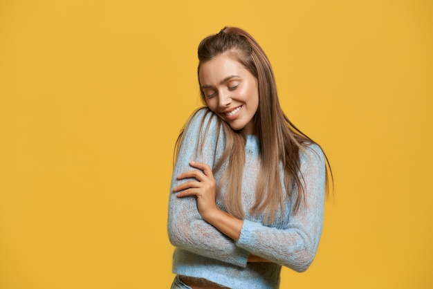 Front view of beautiful young woman with crossed hands closing eyes and grinning. Cute joyful lady pressing cheek to shoulder. Isolated on yellow  wall. Concept of comfort and contentment.
