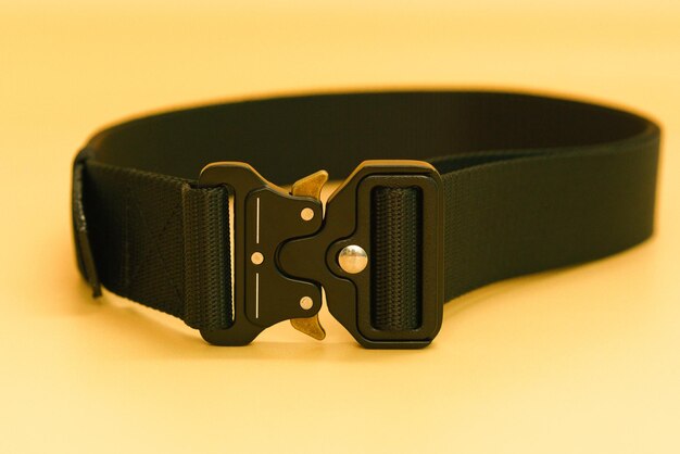 A front view of a black canvas belt with a black metal male\
buckle and a black belt loop lays on the orange background utility\
hiking material outfit safety soldier steel uniform weapon