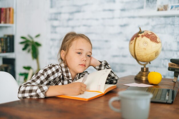 Front view of bored tired cute elementary child school girl doing writing homework holding pen