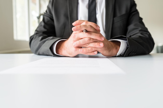 Front view of businessman sitting at his office desk with his hands crossed and a ball point looking out between his fingers.