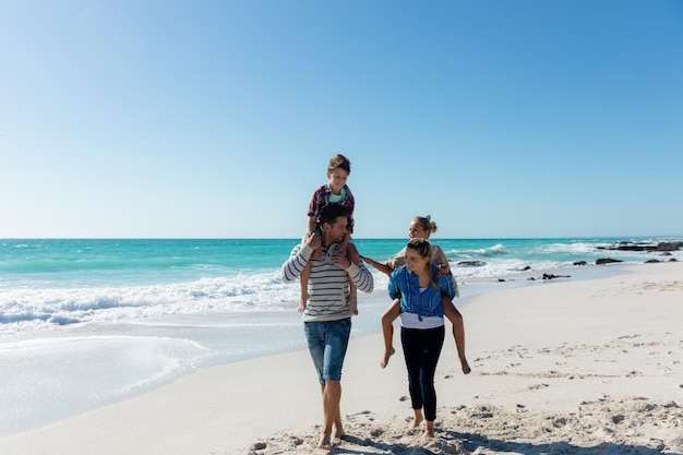 Front view of a Caucasian family on the beach with blue sky and sea in the background, walking, piggybacking and smiling