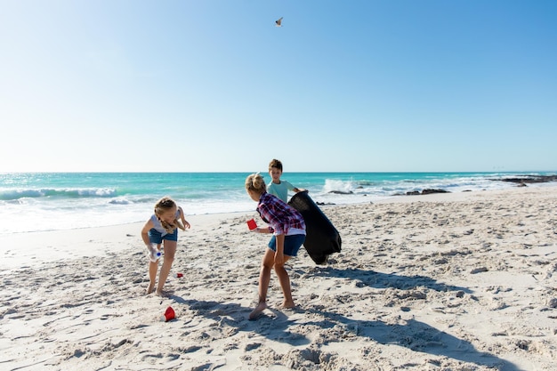 Front view of Caucasian siblings on the beach with blue sky and sea in the background, picking up the rubbish and putting it into a trash bag