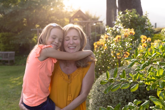 Front view of a Caucasian woman and her granddaughter in the garden on a sunny day, the woman carrying her granddaughter who is embracing her, both are smiling with their eyes closed