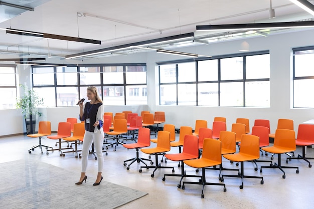 Vista frontale di una donna caucasica con lunghi capelli biondi, che indossa abiti eleganti, in piedi in una sala riunioni moderna e vuota, mentre prepara il suo discorso per la conferenza.