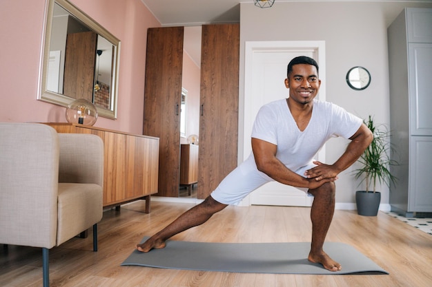 Front view of cheerful African-American man doing side lunge exercise at home during working out standing on yoga mat at bright domestic room, looking at camera . Concept of sport training at home gym