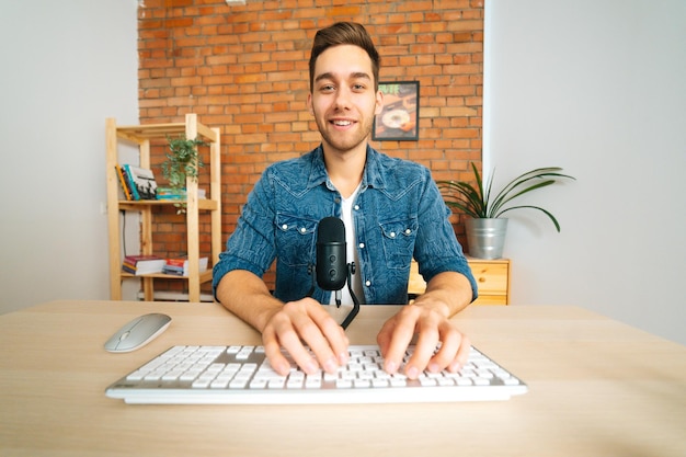 Вид спереди веселого блогера-мужчины, сидящего за столом и говорящего на профессиональном микрофоне, вещающем в домашнем офисе