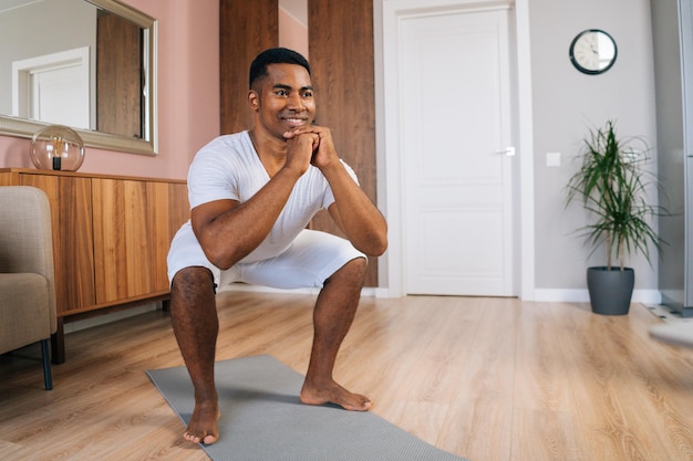 Front view of cheerful muscular African-American man wearing white T-shirt exercising and doing squats online in front of laptop at bright domestic room. Concept of sport training at home gym.