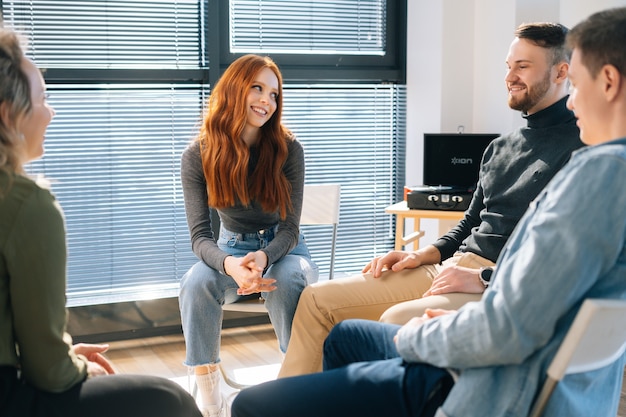 Front view of cheerful young redhead businesswoman talking and discussing new ideas with creative business team, during brainstorming of start-up projects in modern office room near window.