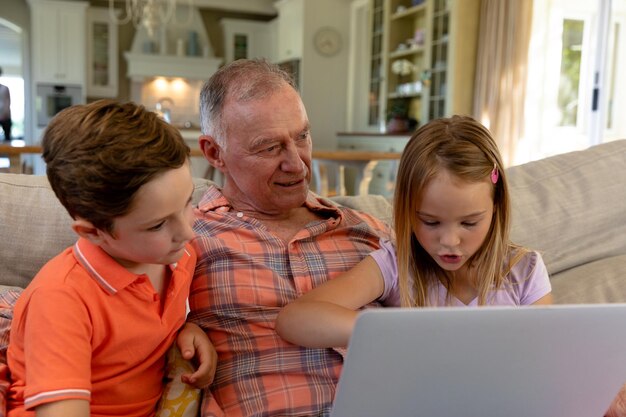 Photo front view close up of a senior caucasian man at home in the living room sitting on the couch with his grandson and granddaughter, using a laptop computer together